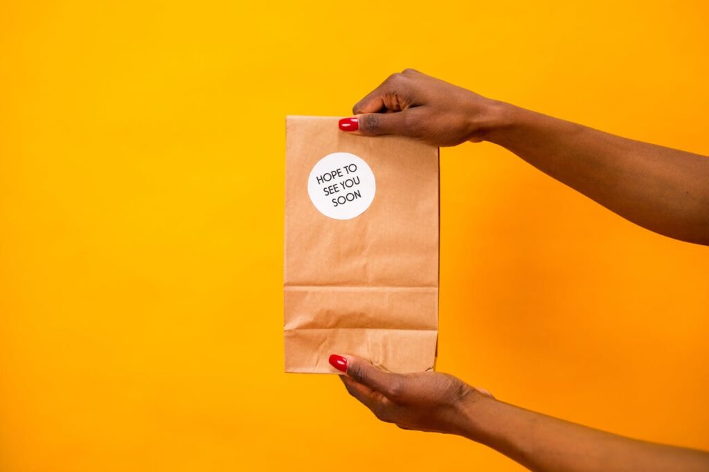 A close-up shot of hands holding a paper bag with 'Hope to See You Soon' text on a vibrant yellow background.